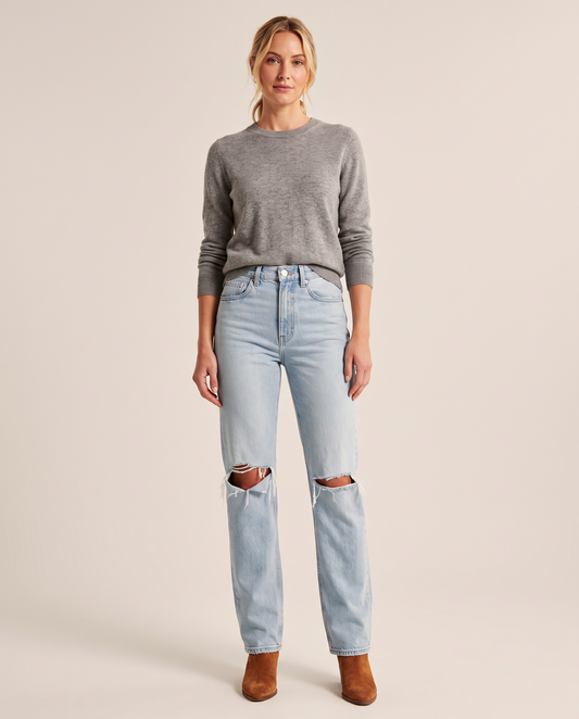 Woman wearing light blue high-waisted straight jeans with ripped knees, a grey merino sweater and cognac suede ankle boots, studio front