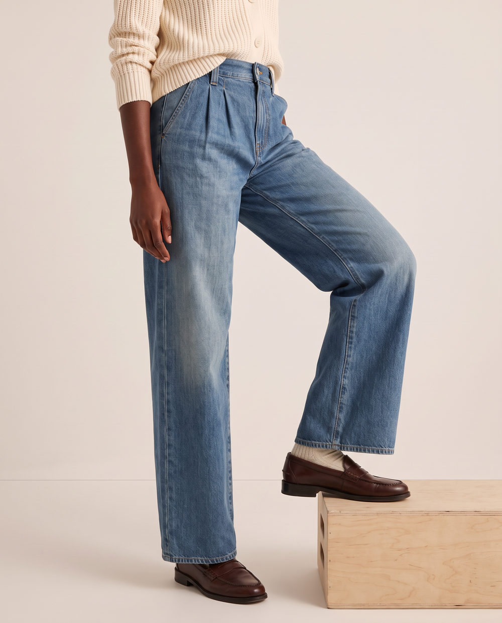Pleated wide-leg jeans in medium blue, waist-down three-quarter view stepping onto a wooden block with brown loafers