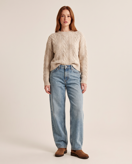 Woman wearing baggy light wash jeans with an oatmeal cable-knit sweater and brown Chelsea boots, studio front view