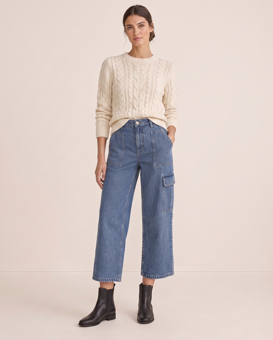 Woman wearing blue cropped cargo jeans with a cream cable-knit sweater and black ankle boots, front view