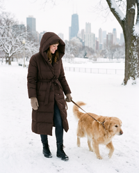 Women's Long Belted Puffer Coat in chocolate brown worn hood up while walking a dog in a snowy Chicago park