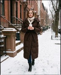 Women's Long Belted Puffer Coat in chocolate brown worn on a snowy New York brownstone street